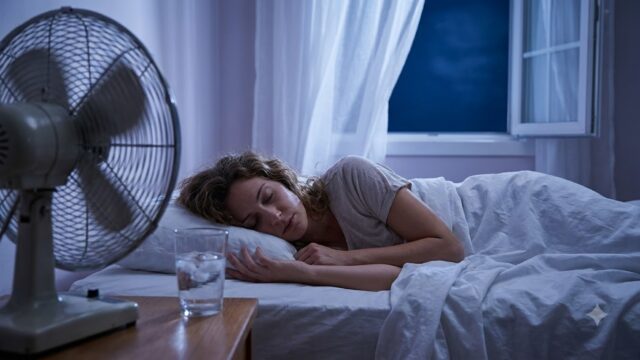 Woman sleeping on light cotton sheets with a fan blowing toward her and an open window on a summer night